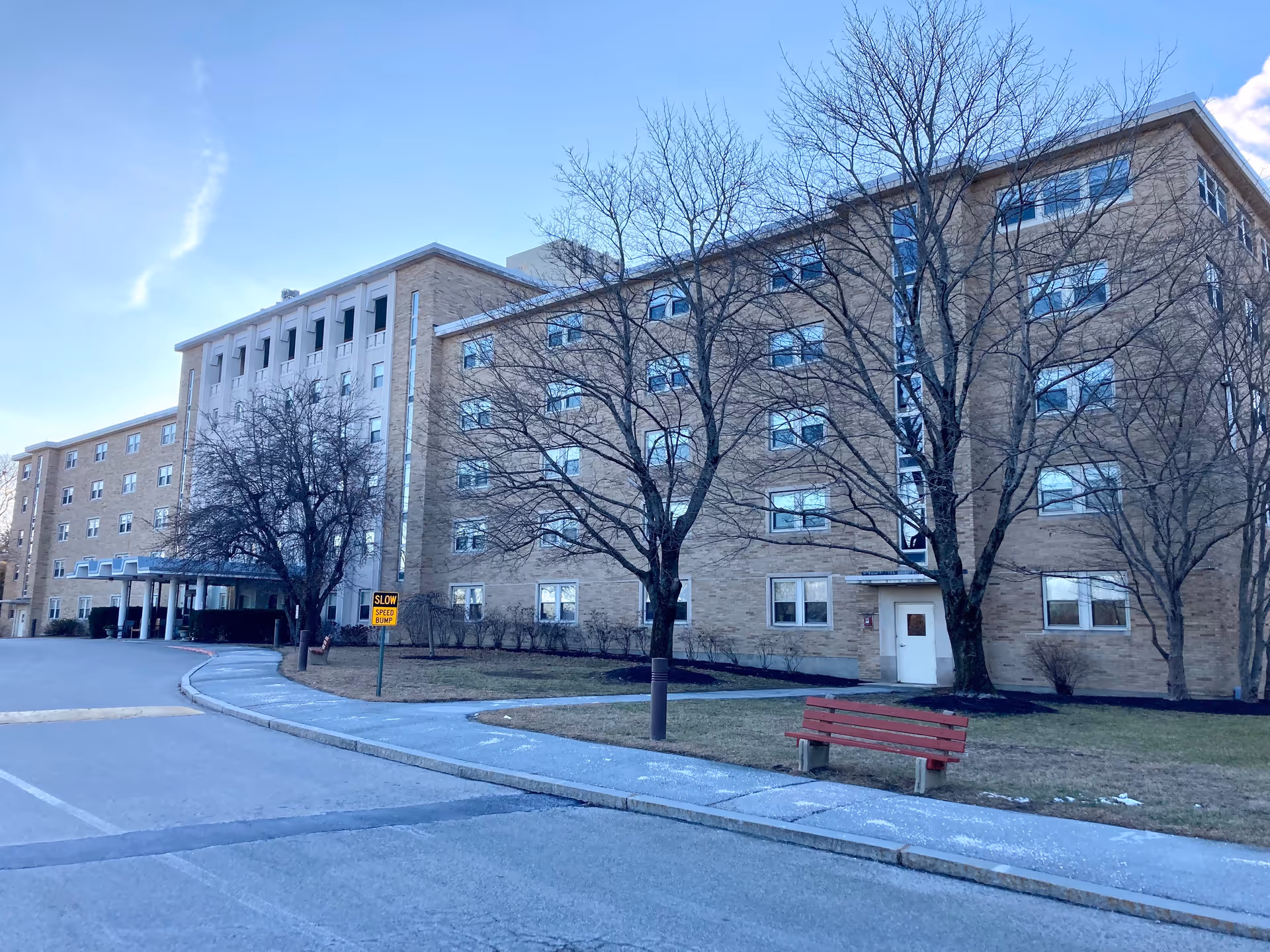 Exterior view of a multi-story brick building with several windows, leafless trees, a red bench on a grassy area, a sidewalk, and a driveway with a yellow 'Slow Speed Bump' sign under a clear blue sky.