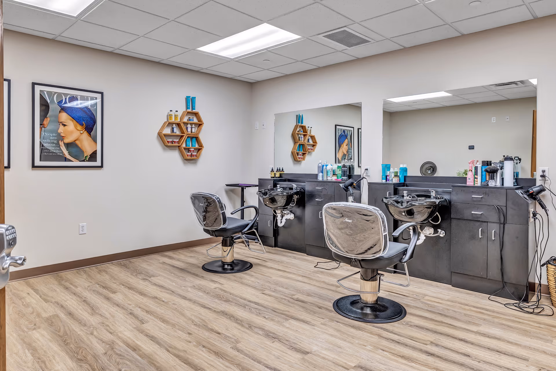 Interior view of a hair salon area in a senior living facility with two black salon chairs in front of a long counter with sinks, mirrors, and various hair care products. The room has light wood flooring, white walls, and a framed Vogue magazine cover on the wall.
