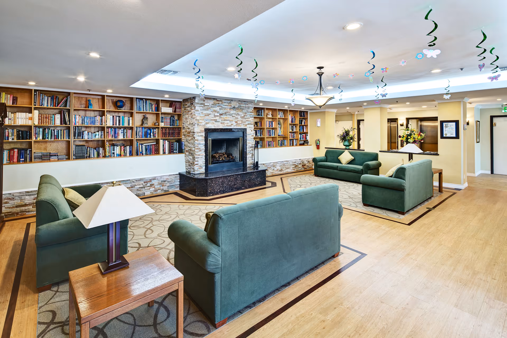 A spacious and well-lit common living area with green upholstered sofas arranged around a central stone fireplace. The room features built-in wooden bookshelves filled with books, a wooden side table with a lamp, and decorative ceiling hangings. The floor has a combination of wood and patterned carpet, and there are flowers and framed pictures on the walls.