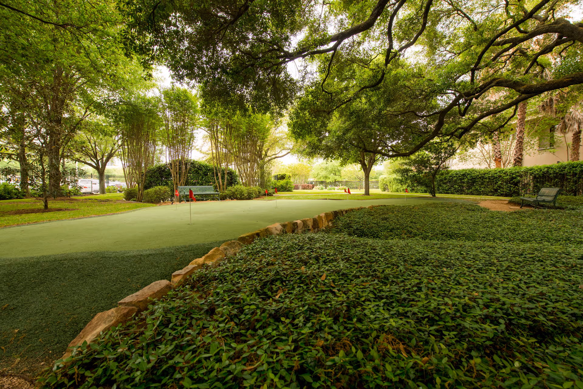 A serene outdoor garden area with lush green trees and bushes surrounding a putting green with small red flags. There are benches placed around the garden for seating, and the scene is shaded by large tree branches overhead.