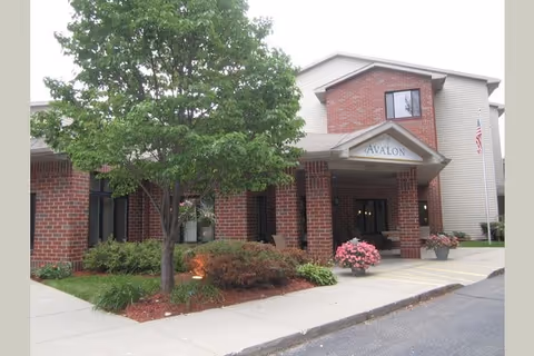 Front entrance of a brick senior living building with a covered portico labeled "Avalon", a tree, and potted flowers.