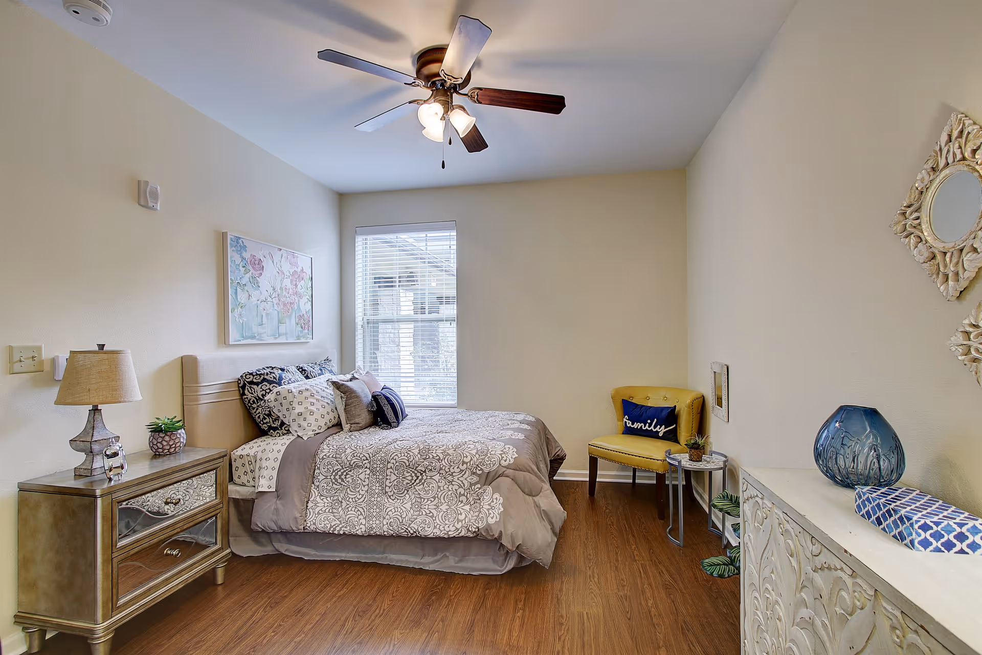 A cozy bedroom with a bed covered in patterned bedding and multiple pillows. There is a wooden nightstand with a lamp and a small plant on it next to the bed. A window with blinds is behind the bed, letting in natural light. A yellow chair with a blue pillow that says 'family' sits in the corner next to a small round table with a plant. On the right side, there is a white ornate cabinet with decorative items including a blue vase and a patterned box. The room has wooden flooring and a ceiling fan with lights.