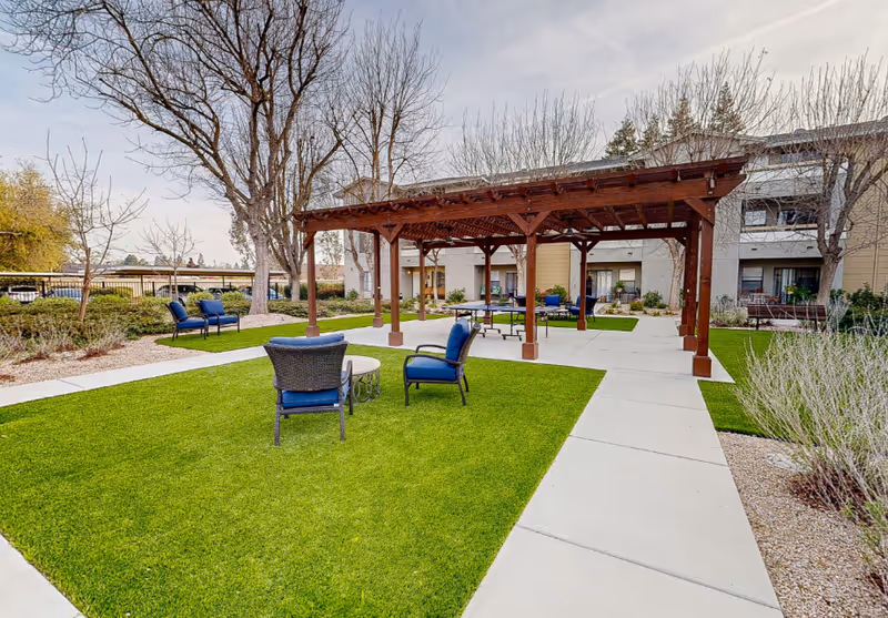 Outdoor courtyard featuring a wooden pergola, blue cushioned patio chairs, concrete walkways and lawn in front of a multi-story senior living building.