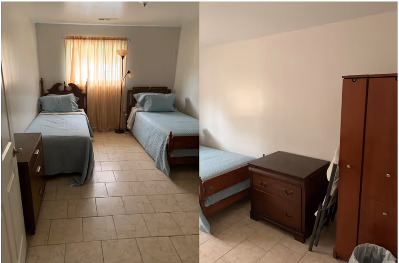 A small bedroom with two single beds covered in light blue bedspreads. There is a window with beige curtains between the beds. A floor lamp stands near the window. The room has tiled flooring and light-colored walls. There is a wooden dresser and a wooden wardrobe along with a folded chair and a trash can.