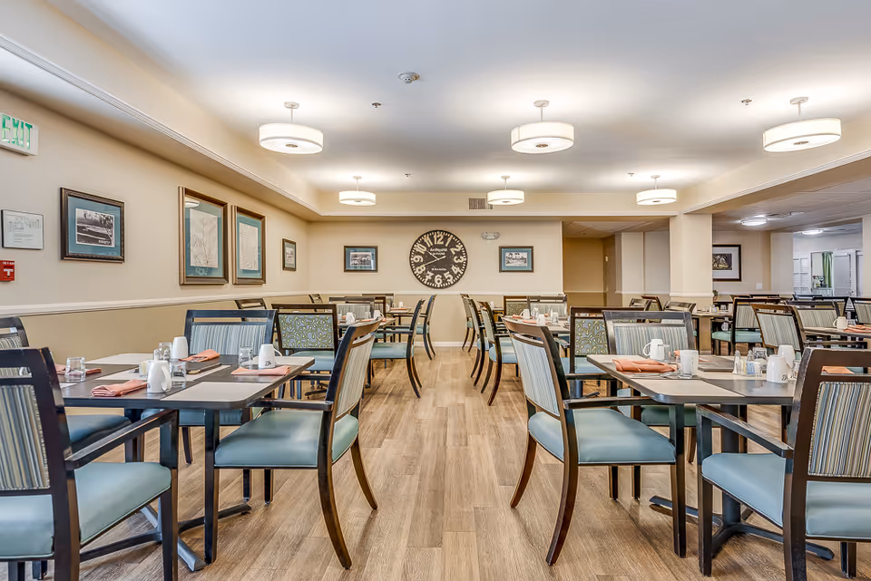 A spacious dining room with multiple tables and chairs arranged neatly. Each table is set with placemats, napkins, cups, and condiments. The room has light-colored walls adorned with framed pictures and a large decorative clock on the far wall. The floor is wooden, and the ceiling has several modern round light fixtures providing bright illumination.