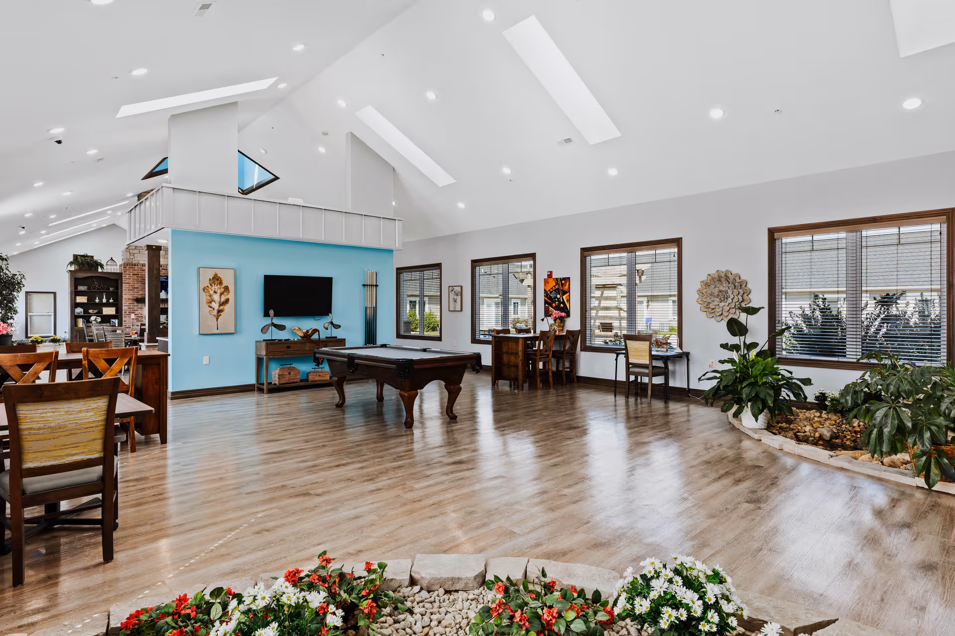 Spacious bright activity room with a pool table, seating areas, indoor plants and large windows under skylights.