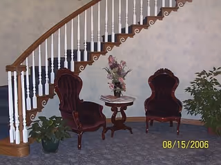 A cozy interior corner featuring two vintage-style burgundy armchairs with wooden frames, a small round wooden table between them holding a floral arrangement, a potted plant on the floor, and a staircase with white balusters and a wooden handrail in the background.