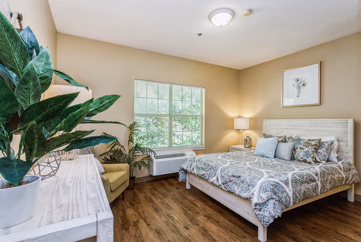 A cozy bedroom with a large window letting in natural light. The room features a wooden bed with patterned bedding and multiple pillows, a nightstand with a lamp and decorative lantern, a framed botanical print on the wall, a wooden dresser with a potted plant, and an armchair next to another plant. The floor is wooden and the walls are painted beige.