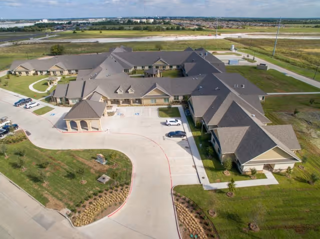 Aerial view of Legend Oaks Healthcare and Rehabilitation - Waxahachie, showing a large, single-story building with multiple wings, surrounded by green lawns and parking areas. The building has a gray roof and beige exterior walls, with a covered entrance and several cars parked in front.