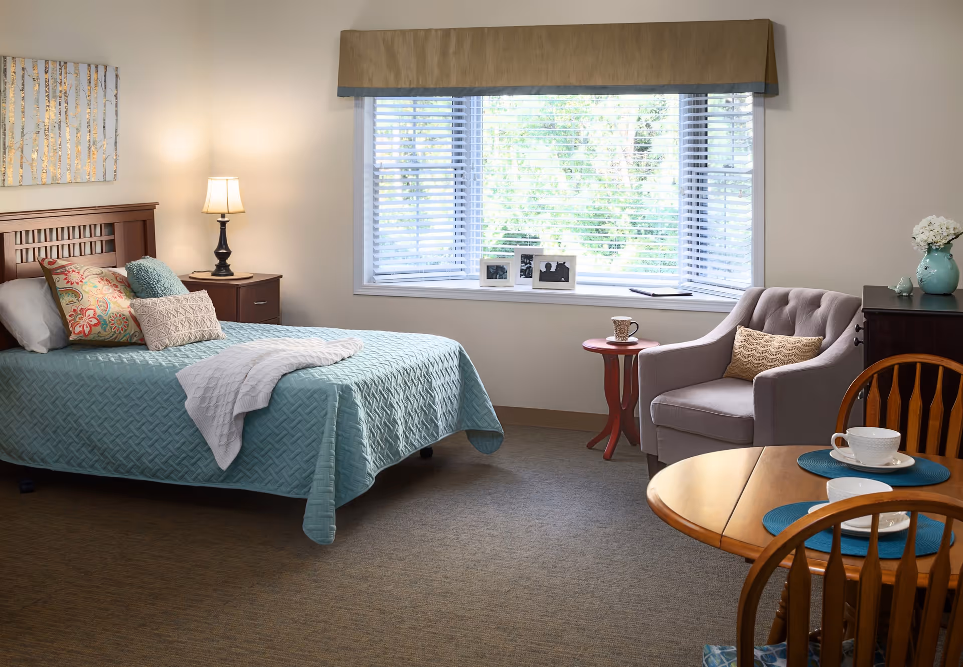 A cozy bedroom with a bed covered in a light blue quilt and multiple decorative pillows. Next to the bed is a wooden nightstand with a lamp. A large window with blinds lets in natural light, and a small red side table holds a cup. There is a comfortable gray armchair with a patterned pillow, a wooden dining table with chairs, and a vase with white flowers on a dark wooden dresser.