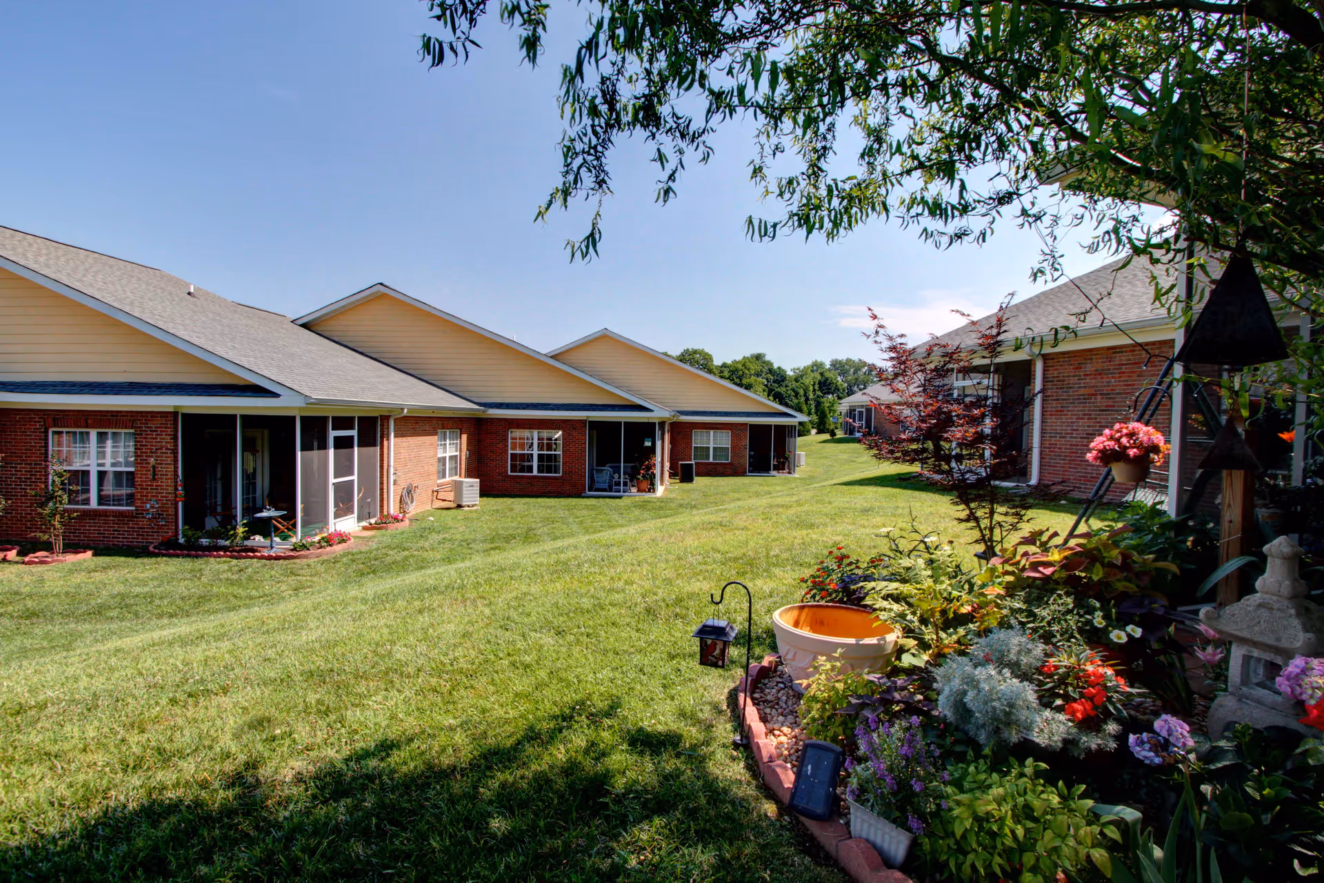 View of a grassy outdoor area between single-story brick buildings with beige roofs at a senior living facility. The foreground features a well-maintained garden with various colorful flowers and plants, and a small decorative lantern. The sky is clear and blue.