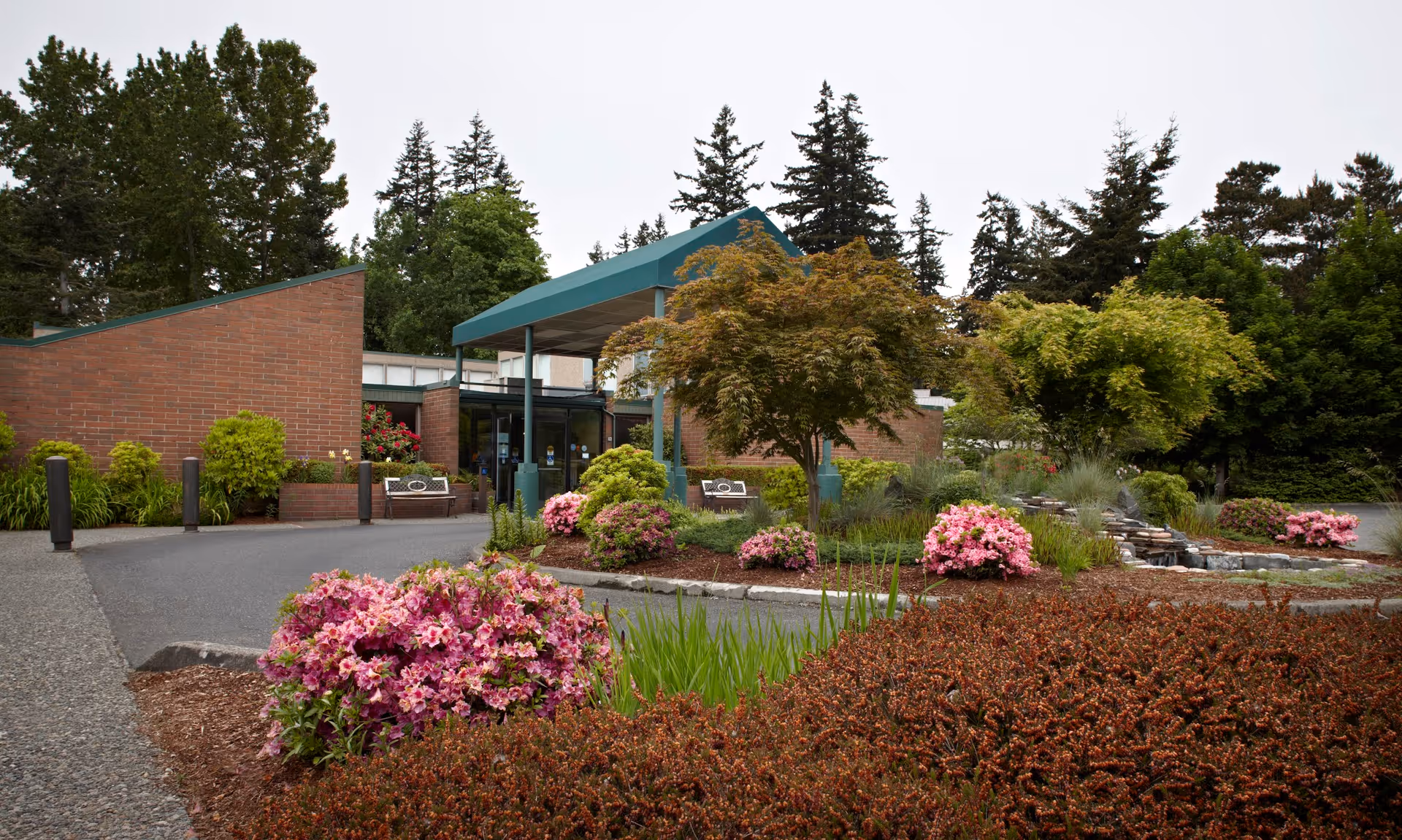 Entrance of Avamere Bellingham Healthcare and Rehabilitation Services with a covered drop-off area, surrounded by landscaped gardens featuring various bushes, flowering plants, and trees.