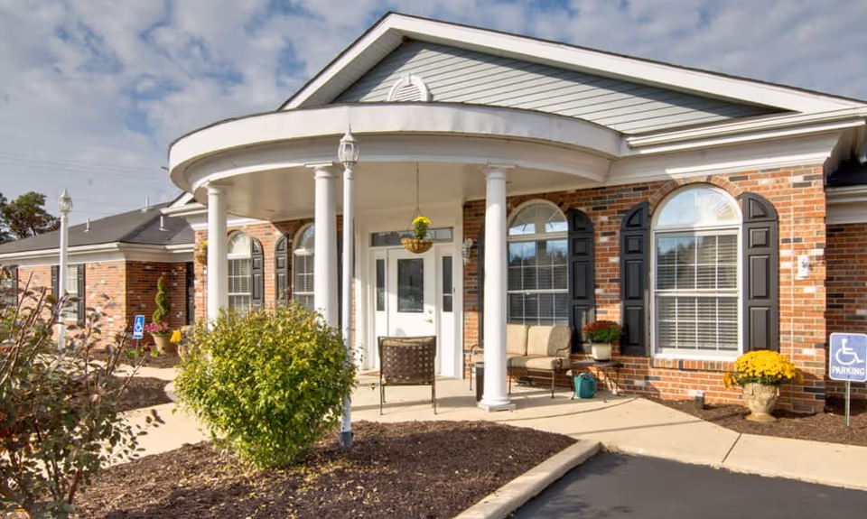 Front exterior view of a brick building with white columns supporting a curved porch roof. There are large windows with black shutters, a white door, outdoor seating with chairs and a bench, potted plants, and a handicapped parking sign visible near the entrance.