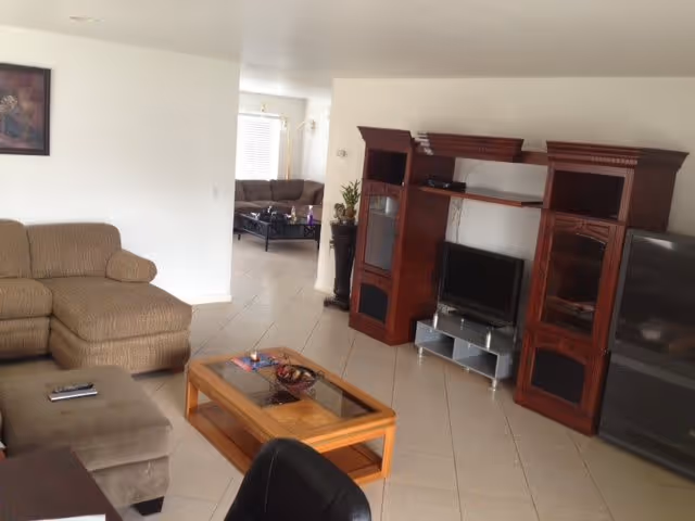 A living room with beige sectional sofa and ottoman, a wooden coffee table with glass top, a dark wooden entertainment center with a flat-screen TV, and a large TV on the right side. The room has tiled flooring and white walls. In the background, there is another seating area with a brown sofa and a coffee table near a window with blinds.