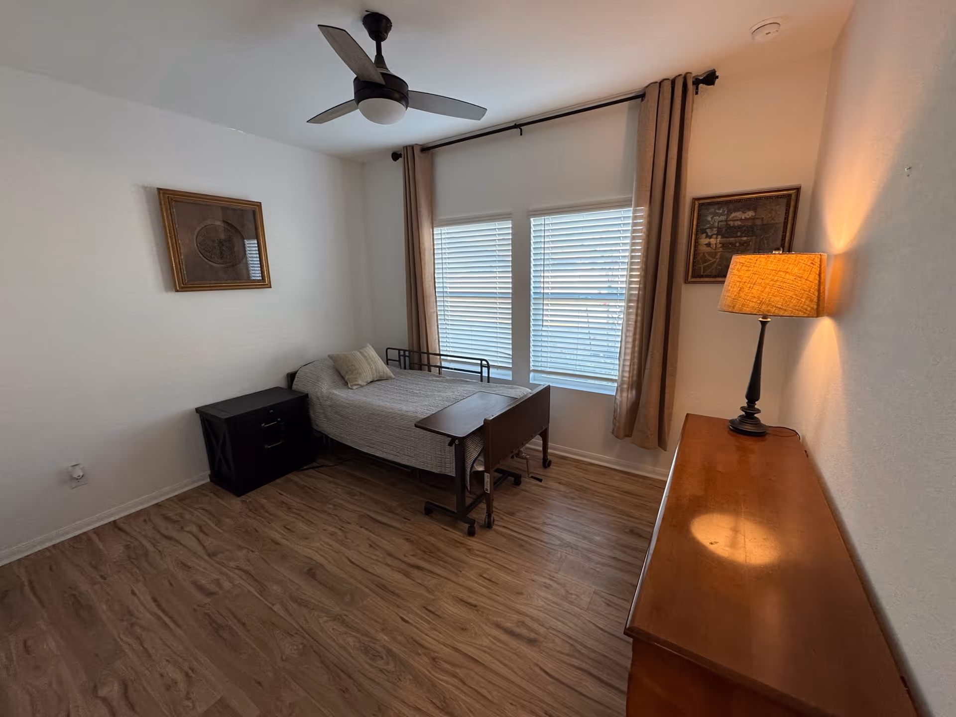 A simple bedroom with a single bed covered with a gray bedspread and a pillow. There is a black nightstand next to the bed and a wooden dresser with a lit lamp on top. Two framed pictures hang on the walls, and a ceiling fan is mounted above. The room has wood flooring and two windows with blinds and beige curtains.