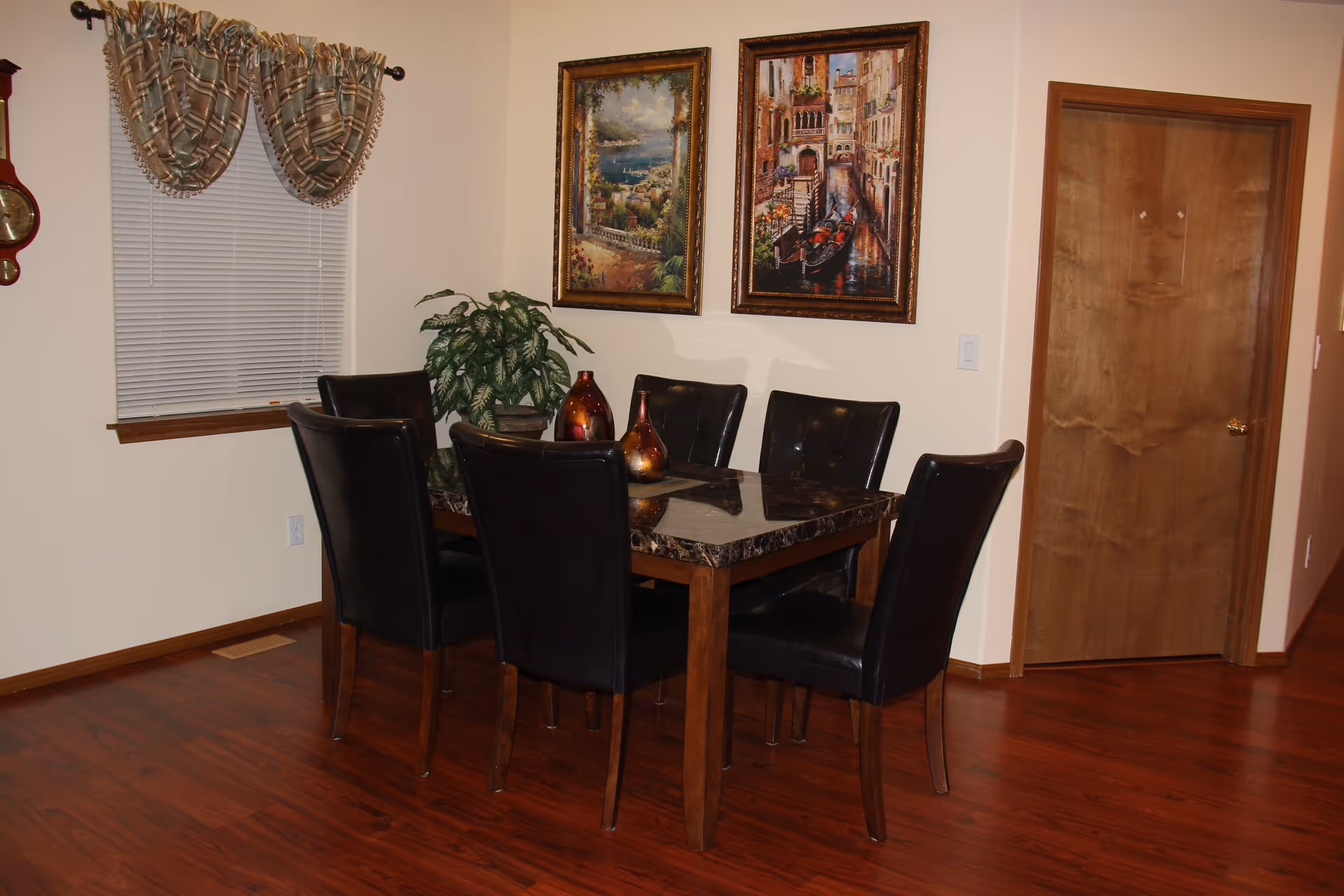 A dining area with a rectangular table featuring a dark marble top and six black leather chairs. Two decorative vases are placed on the table. The room has wooden flooring, a window with blinds and patterned curtains, two framed paintings on the wall, and a wooden door.