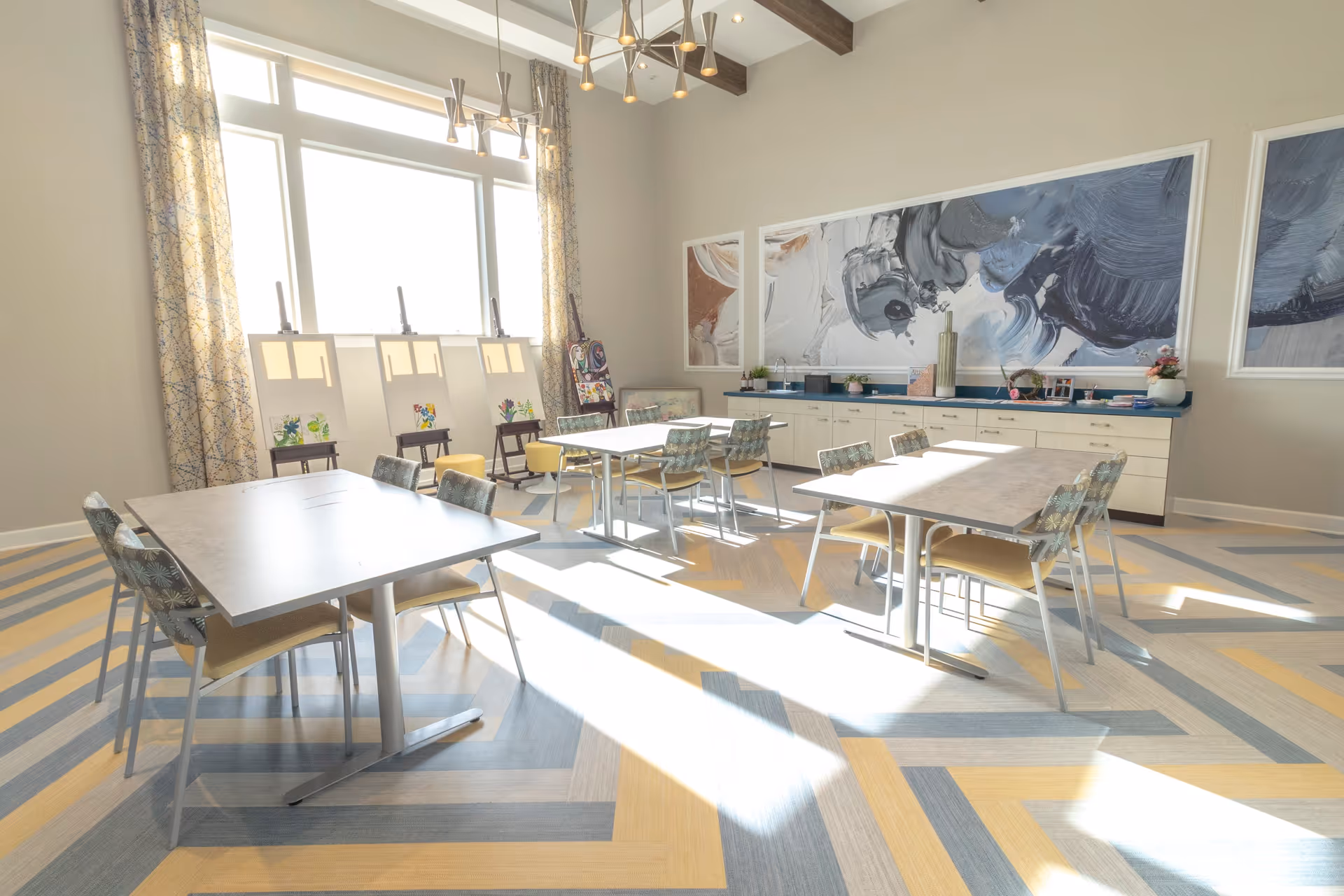 Bright activity room with tables and chairs, easels by a large window, and large abstract artwork on the wall.