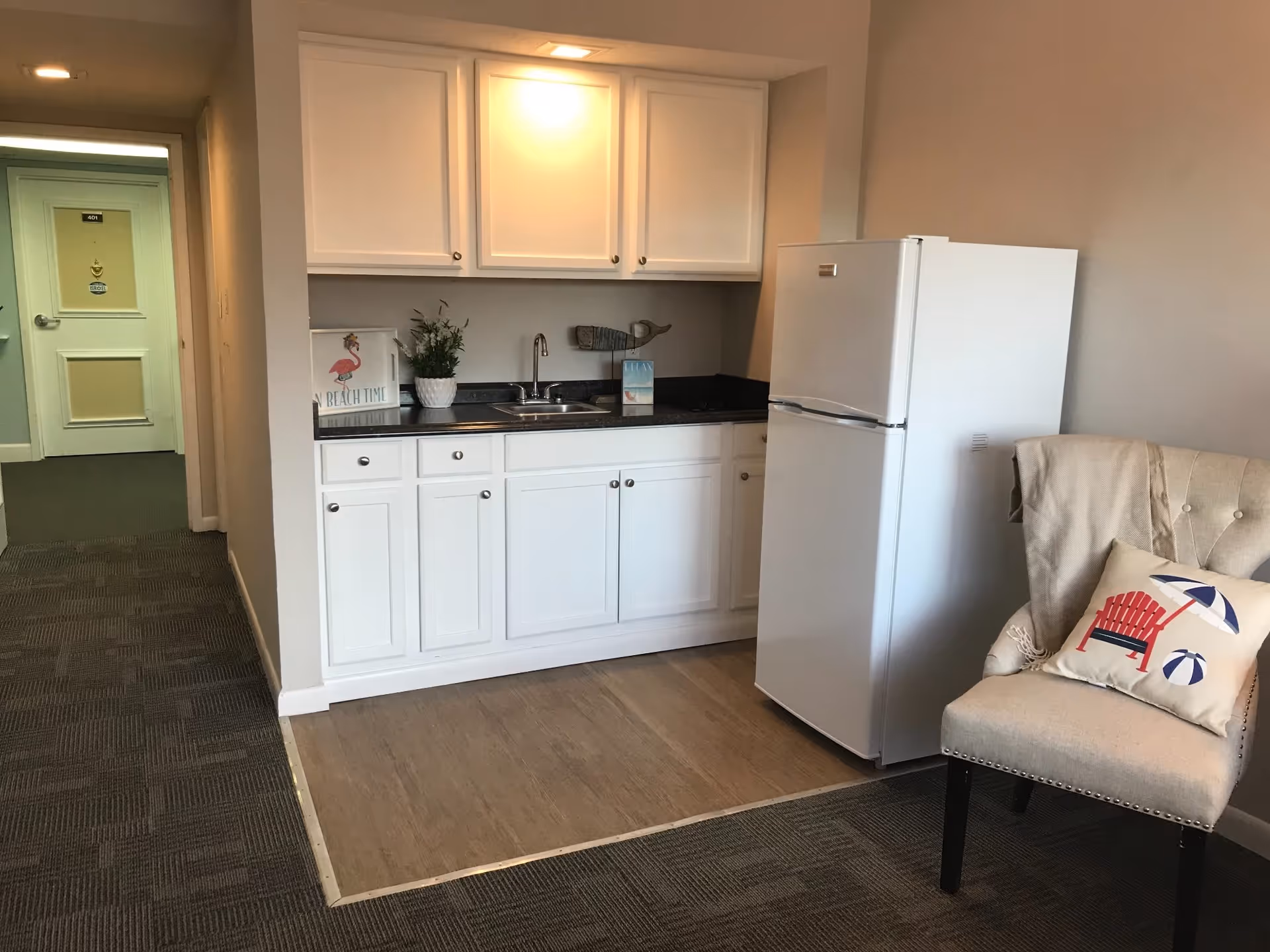 Small kitchenette with white cabinets, a sink, refrigerator and an upholstered chair in a senior living unit.