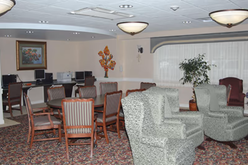 A communal room with a patterned carpet, several upholstered armchairs, a long table surrounded by wooden chairs with striped cushions, and a row of computer stations against the wall. The room is softly lit with ceiling lights and has a large window with sheer curtains and a potted plant nearby.