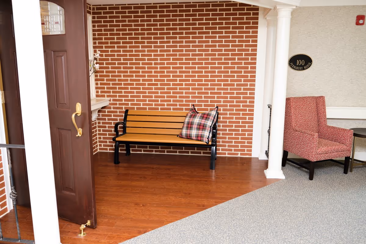 An interior corner of a senior living facility featuring a wooden bench with a plaid cushion against a red brick wall, a partially open dark wooden door with a brass handle, two white columns, and a red upholstered armchair next to a small round table. A plaque on the wall reads '100 PENNSBURY MANOR.'