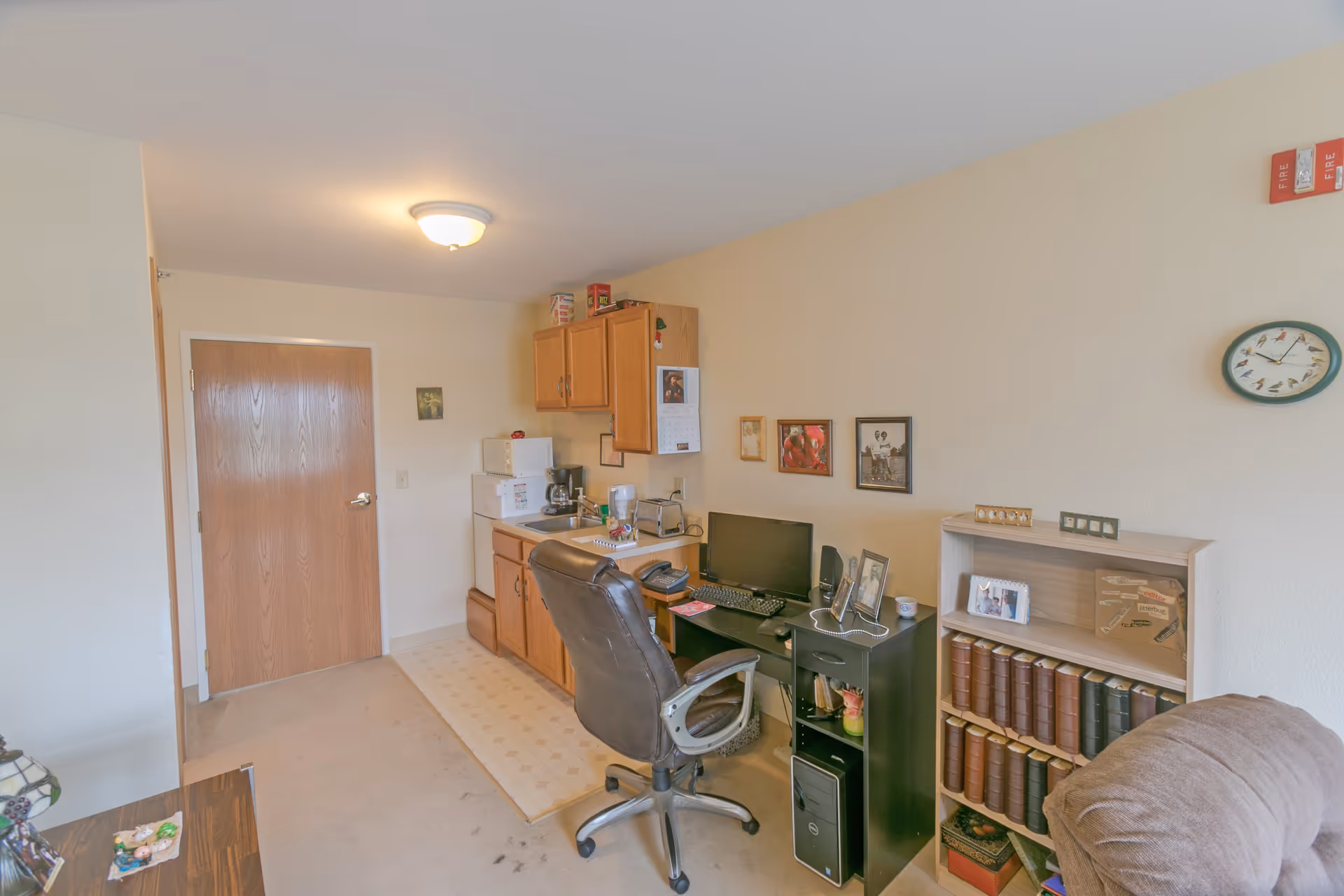 A small room with a wooden door, a kitchenette area with cabinets, a microwave, and a coffee maker. There is a black office chair in front of a desk with a computer monitor, keyboard, and phone. On the right side, there is a bookshelf filled with books and framed photos on the wall above it. The room has beige walls and carpeted floor.