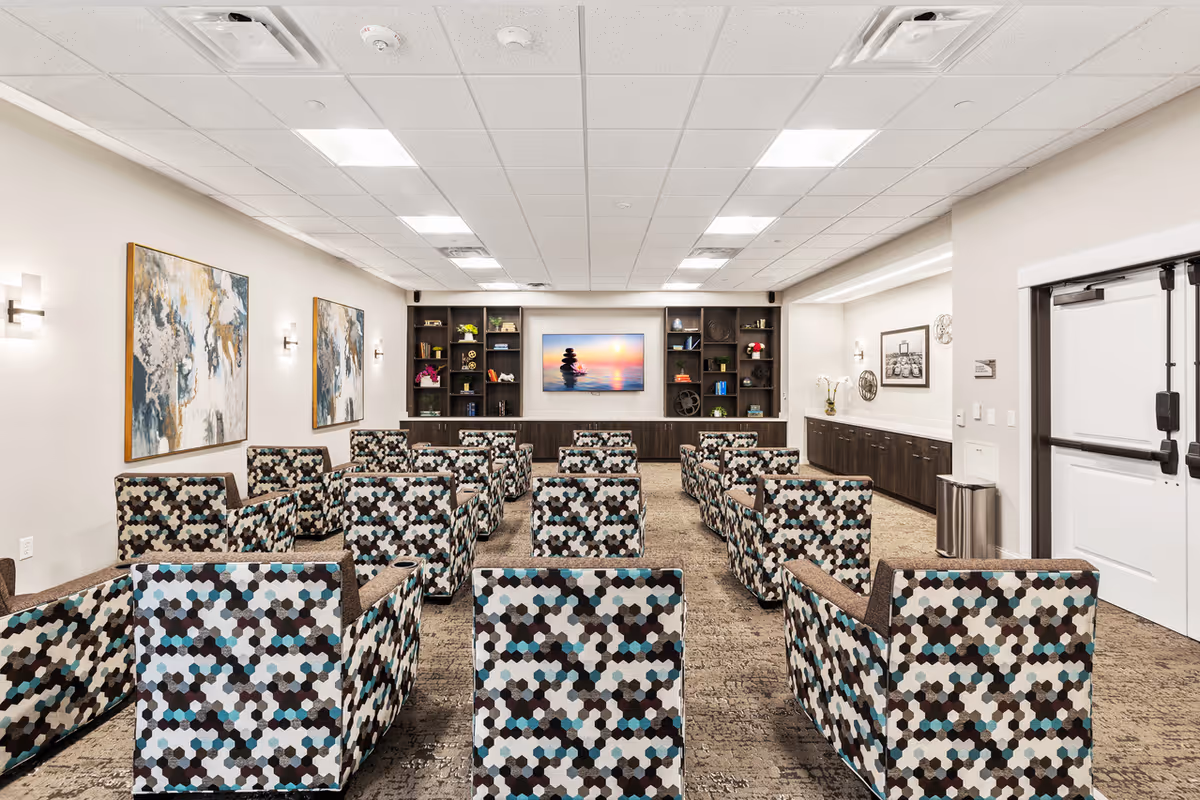 A bright and modern common area with multiple patterned armchairs arranged in rows facing a large flat-screen TV mounted on a wall with built-in shelves displaying decorative items. The room has a carpeted floor, white walls with abstract paintings, and recessed ceiling lights. There is a double door on the right side and a long cabinet with additional decor on the right wall.