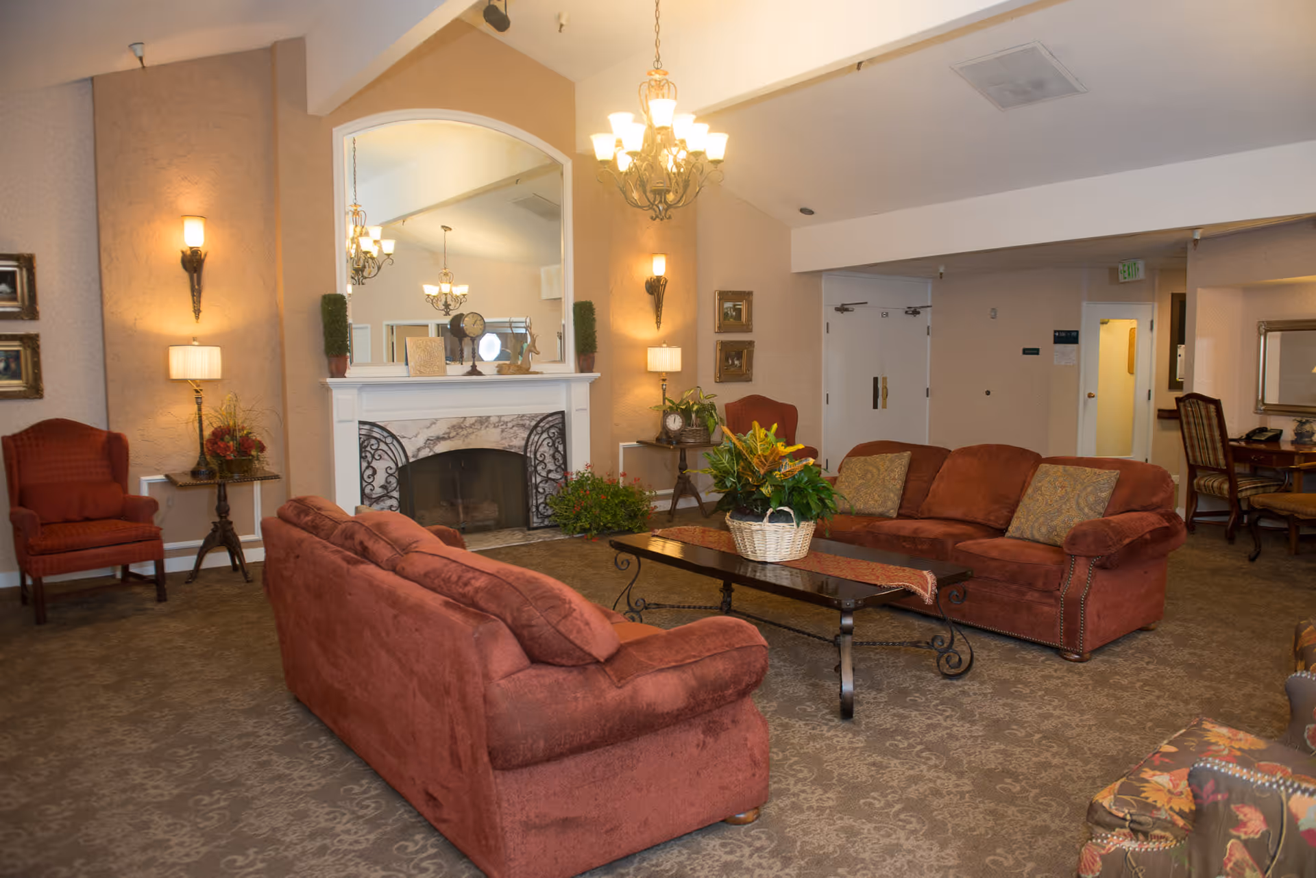 A cozy living room area in a senior living facility with two red sofas facing each other around a dark wooden coffee table with a plant centerpiece. The room features a fireplace with a large mirror above it, wall sconces, a chandelier, and additional seating including an armchair and a desk with a chair in the background.