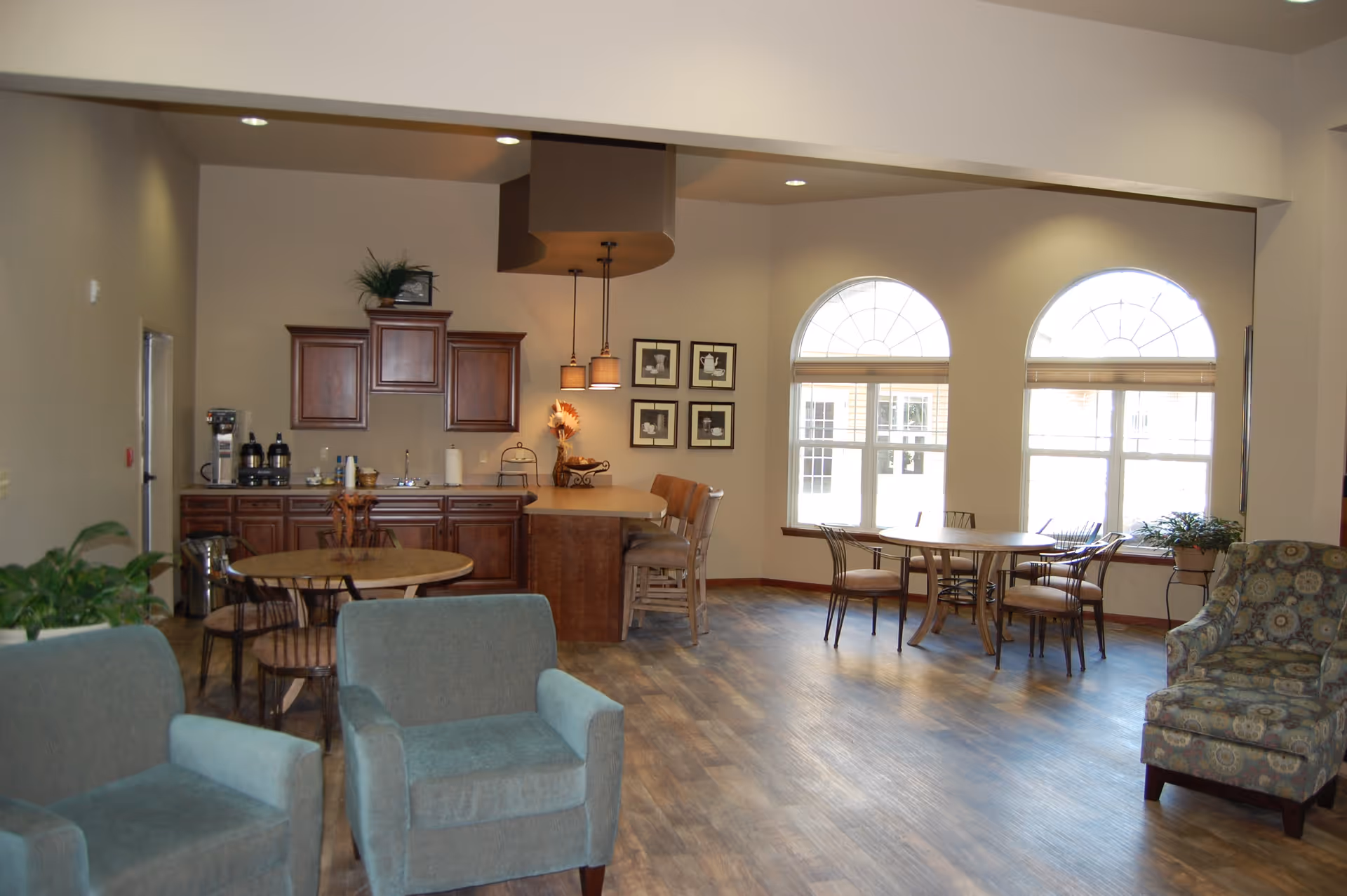 A spacious common area in an assisted living facility featuring a kitchenette with wooden cabinets, a countertop with bar stools, round tables with chairs, two large arched windows letting in natural light, and comfortable upholstered armchairs.