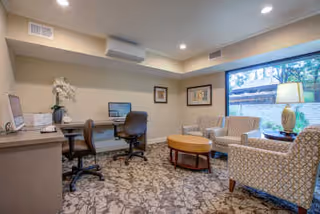 A cozy interior room featuring a workspace with two office chairs and computers on a long desk along one wall. Across from the desk are two patterned armchairs and a matching loveseat arranged around a round wooden coffee table. A large window lets in natural light, and a table lamp sits on a side table next to the loveseat. The room has recessed lighting and a patterned carpet.