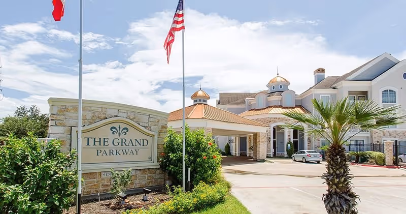 Exterior view of The Grand Parkway senior living facility with a stone sign displaying the facility name, two flagpoles with flags, landscaped greenery, and a driveway leading to the building entrance with a covered drop-off area.