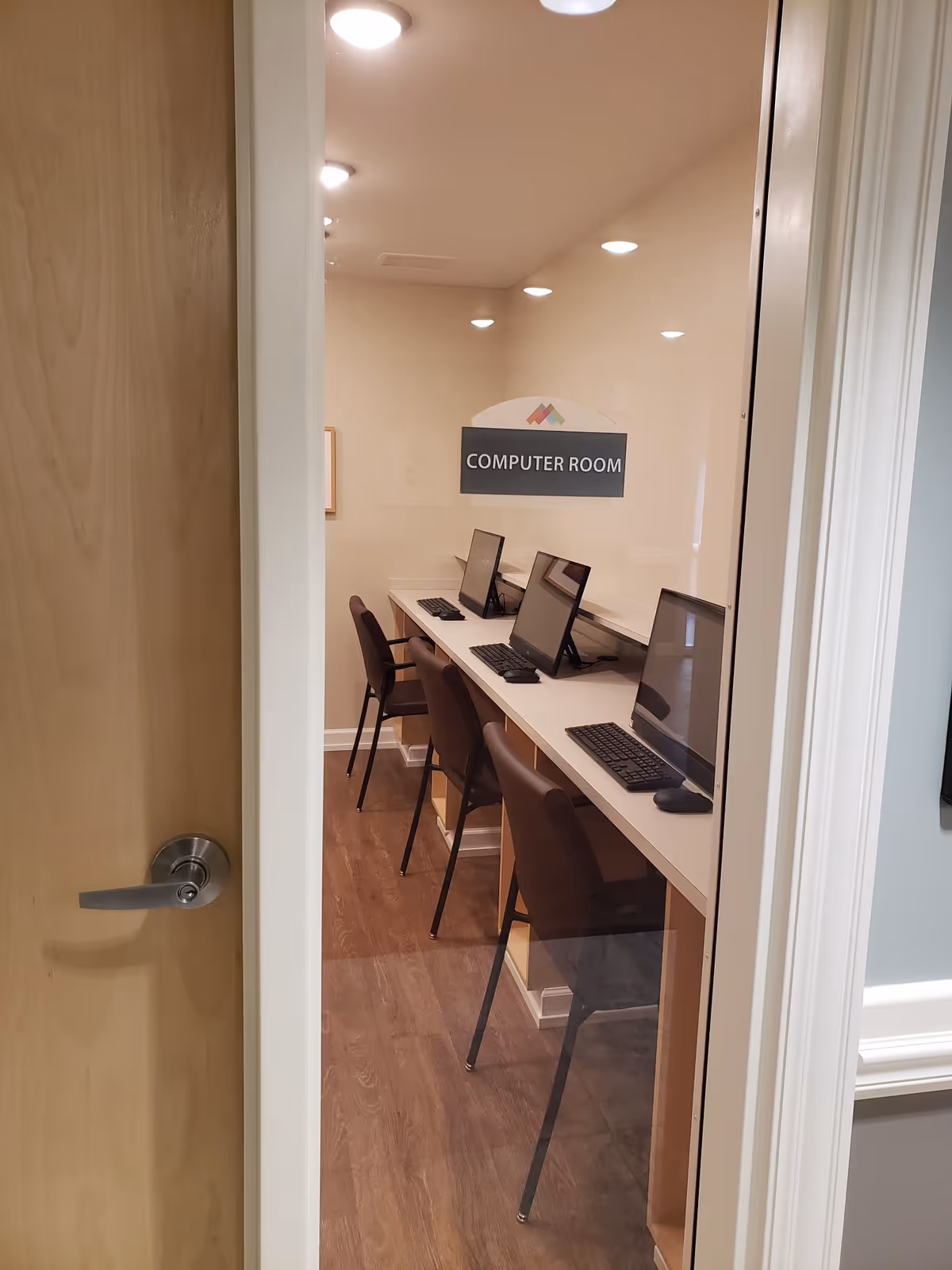 View through a partially open door into a computer room with a row of four computers on a long counter and four chairs. The room has wood flooring and recessed ceiling lights. A sign on the wall reads 'COMPUTER ROOM'.