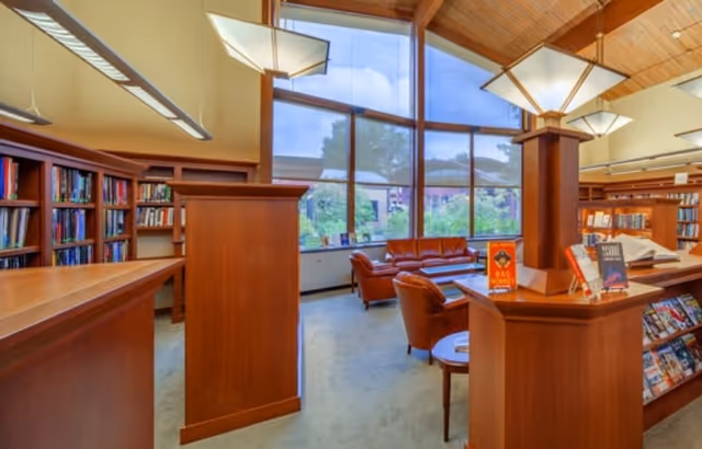 Interior view of a library or reading room with wooden bookshelves filled with books, a seating area with leather chairs and a sofa near large windows that let in natural light, and modern hanging light fixtures from a wooden ceiling.