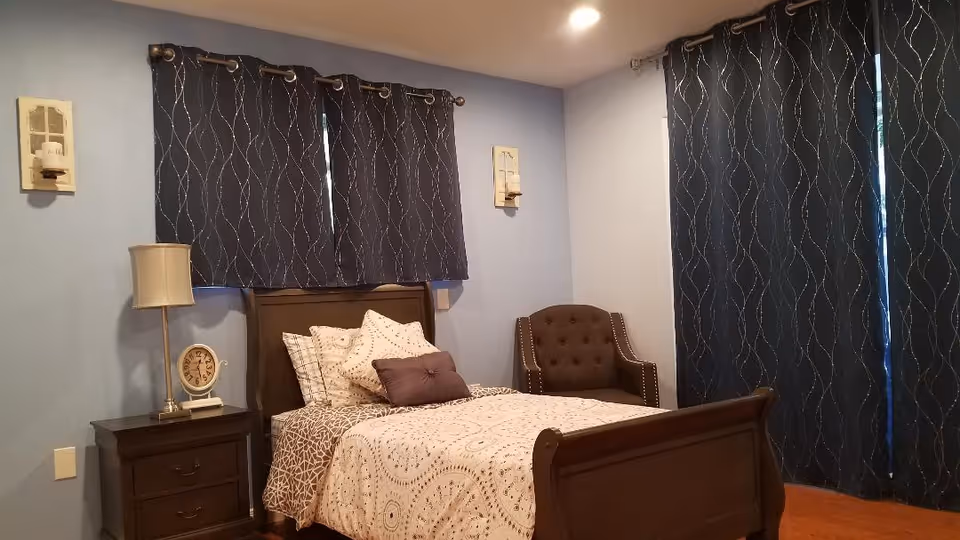 Neat single bedroom with a wooden bed, nightstand with lamp and clock, an upholstered armchair, and dark patterned curtains against blue walls.