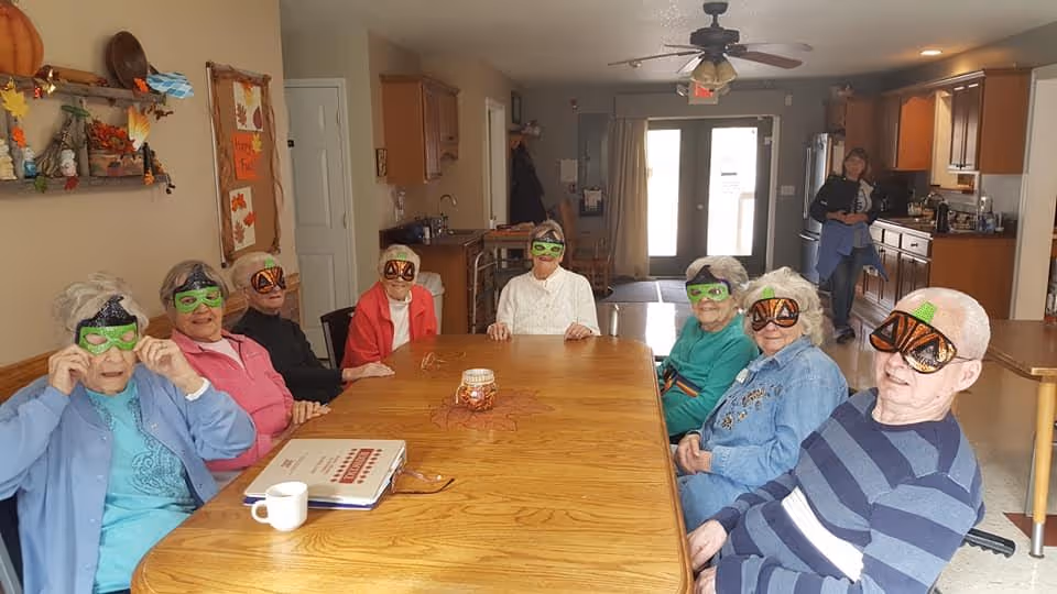 A group of elderly people sitting around a large wooden table in a common area of an assisted living facility. They are wearing festive Halloween-themed glasses and smiling. The room has a ceiling fan, kitchen area in the background, and a staff member standing near the kitchen. The walls are decorated with autumn-themed decorations.