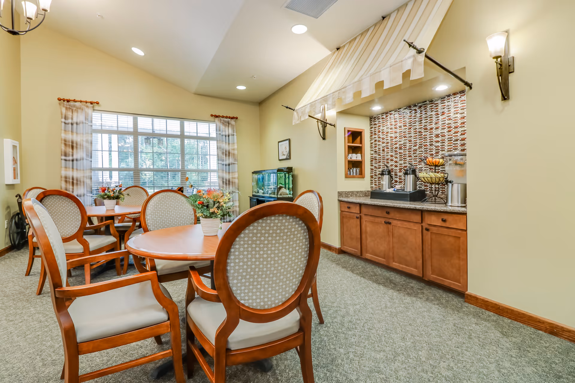 A cozy dining area in an assisted living facility with round wooden tables and cushioned chairs. The room has a large window with patterned curtains allowing natural light to enter. There is a small refreshment counter with coffee dispensers, a water dispenser, and a fruit basket under a striped canopy. A fish tank is visible against the wall, and the walls are painted a soft yellow color.