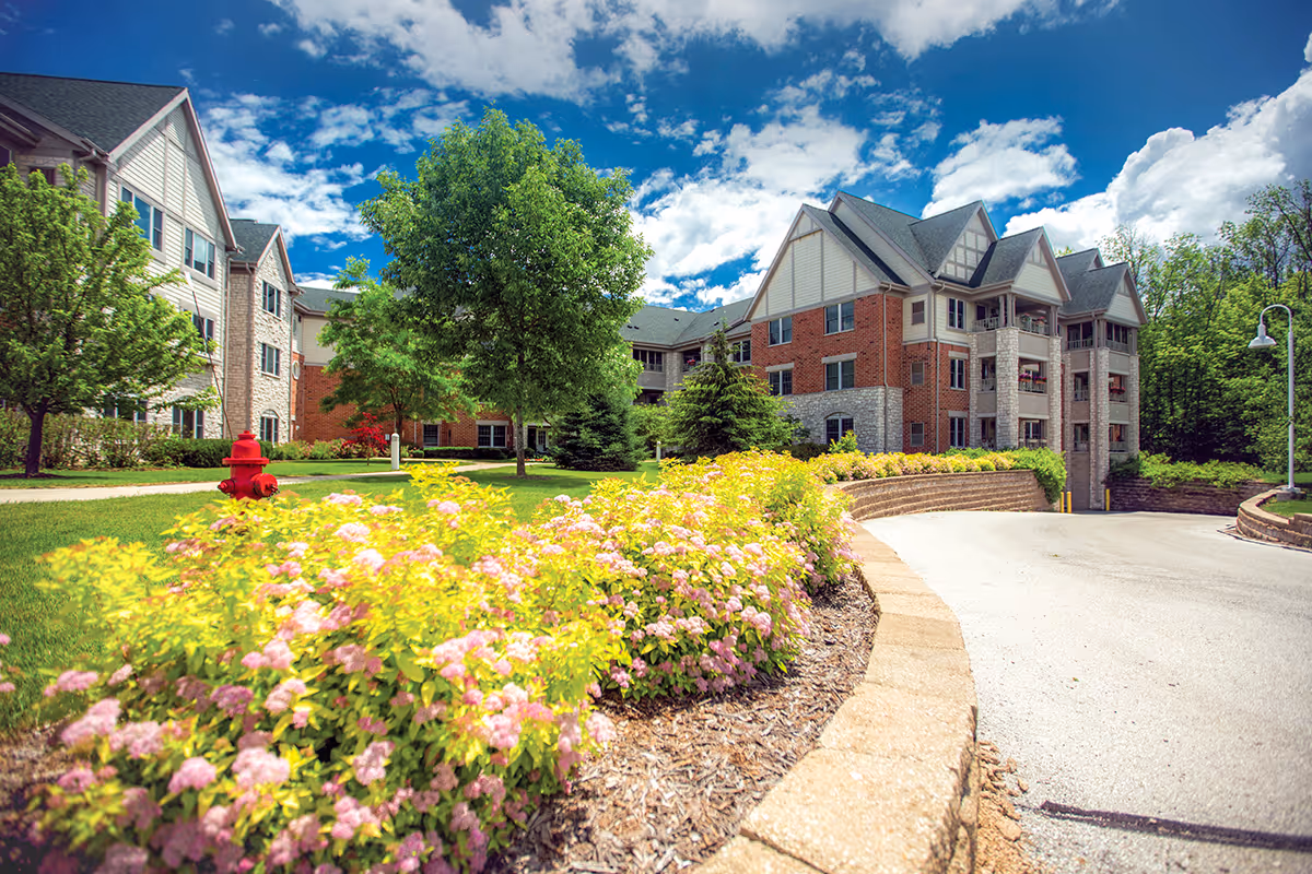 Exterior view of Newcastle Place senior living facility with a curved driveway, landscaped garden featuring pink and yellow flowers, green trees, and a bright blue sky with scattered clouds.