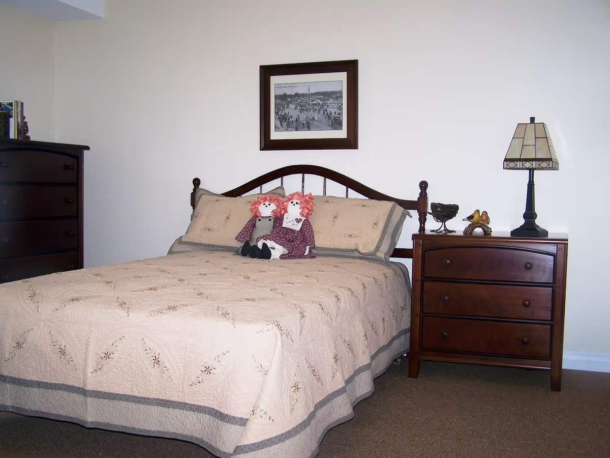 A bedroom with a bed covered in a beige quilt with floral embroidery. Two rag dolls with red hair are sitting on the bed. To the right of the bed is a wooden nightstand with three drawers, a decorative lamp, and small bird figurines. To the left is a tall wooden dresser with books on top. A framed black and white picture hangs on the wall above the bed.