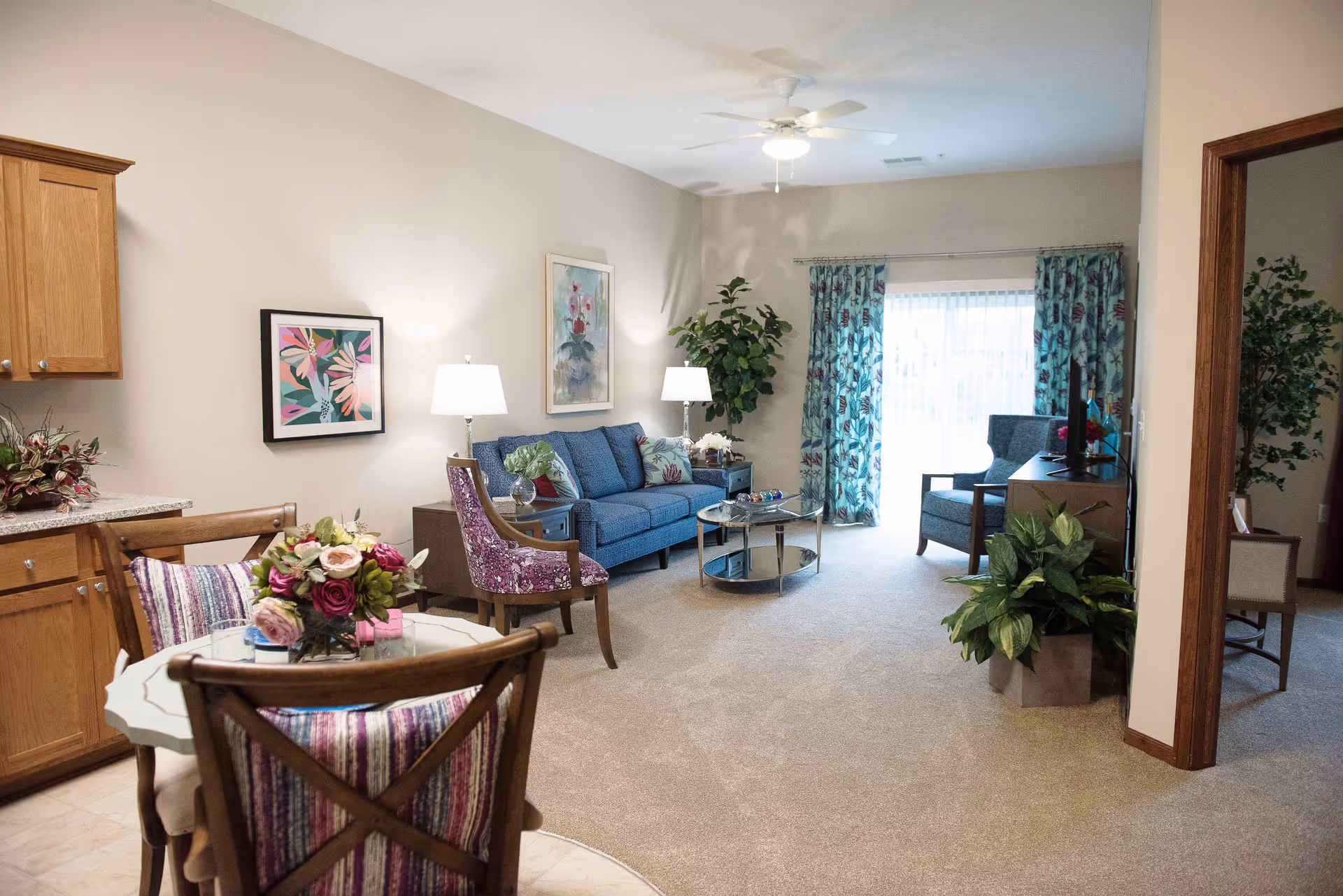 A bright and cozy living room area in a retirement community featuring a blue sofa with patterned cushions, a purple armchair, two side tables with lamps, a round glass coffee table, and a large potted plant near a sliding glass door with floral curtains. Adjacent to the living area is a small dining table with four wooden chairs, decorated with a floral centerpiece. The room has beige carpeting and light-colored walls with framed artwork.
