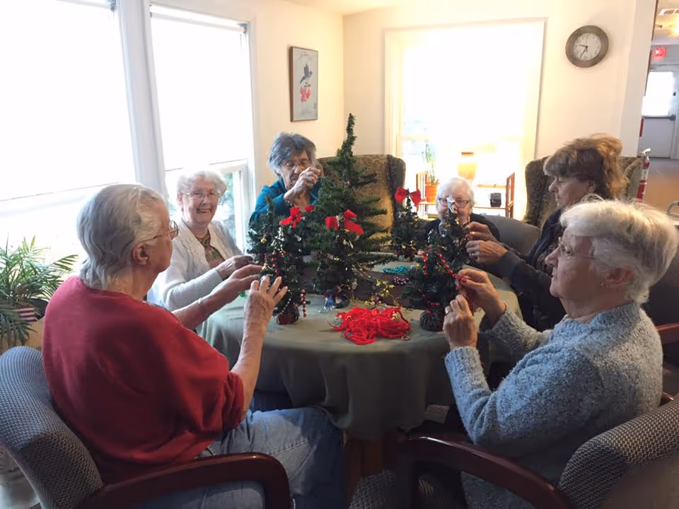 A group of six elderly women sitting around a round table in a well-lit room, decorating small Christmas trees with red ribbons and garlands. The room has large windows with white blinds, a clock on the wall, and comfortable chairs. The atmosphere appears warm and communal.