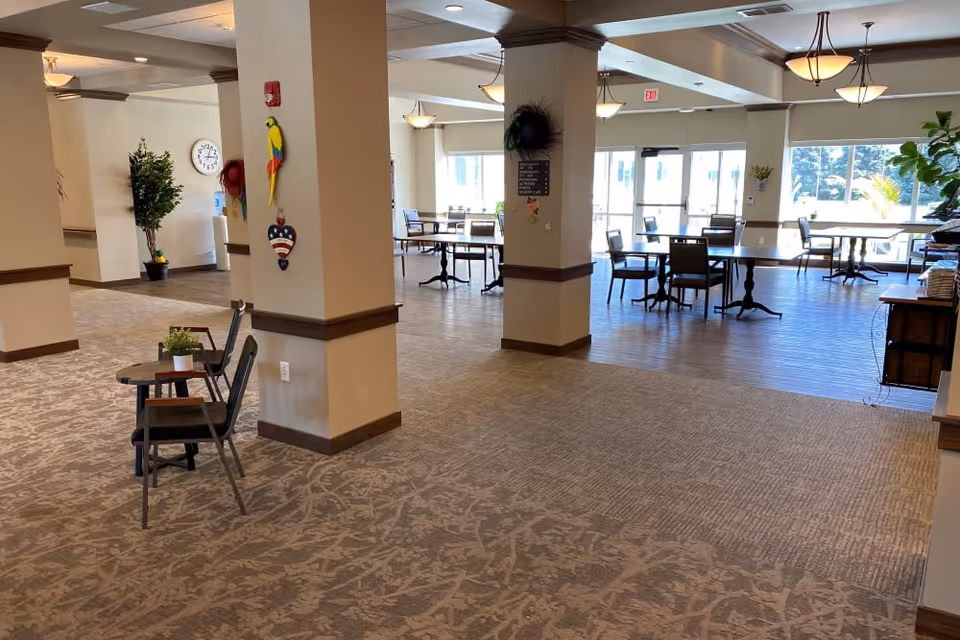 A spacious and well-lit common area in a senior living facility with carpeted flooring and several columns. There are small tables with chairs arranged near large windows that let in natural light. The walls are decorated with colorful wall hangings, and there are plants placed around the room. The area appears clean and inviting with a mix of seating options.