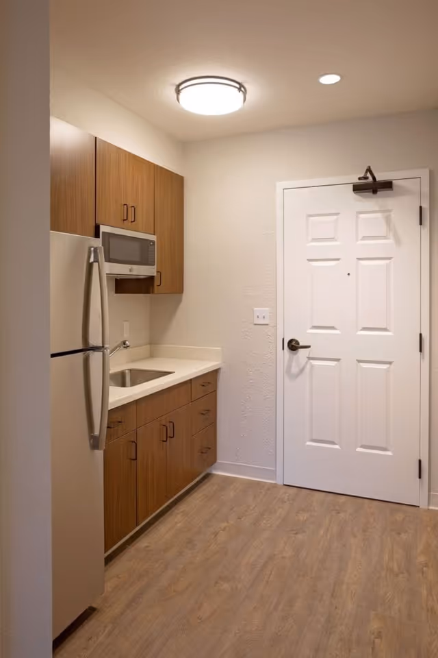 Small kitchen area with wooden cabinets, a stainless steel refrigerator, a microwave mounted above the countertop, a sink, and a white door with a door closer. The floor is wood-patterned and the walls are light-colored.