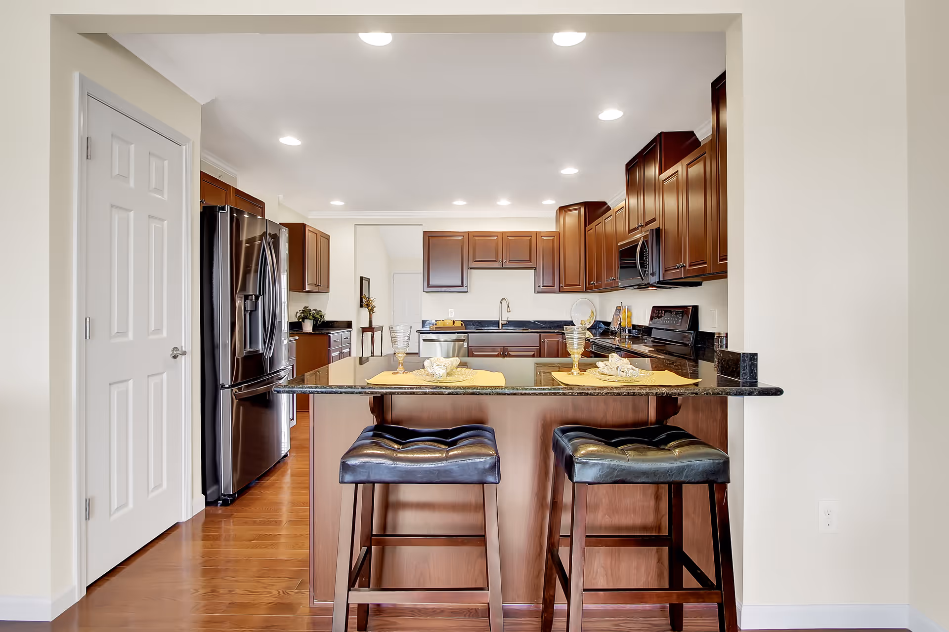 Modern kitchen with dark wood cabinets, black granite countertops, and stainless steel appliances including a refrigerator, microwave, and stove. Two black cushioned bar stools are placed at the kitchen island, which has place settings with yellow placemats and glassware. The kitchen has recessed lighting and hardwood floors.