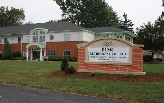 Exterior view of Elms Retirement Village building with a large brick sign in front displaying the facility name and services including skilled nursing, rehabilitation, and assisted living. The building has a combination of red brick and white siding with multiple windows and a green roof over the entrance. There are trees and well-maintained grass surrounding the area.