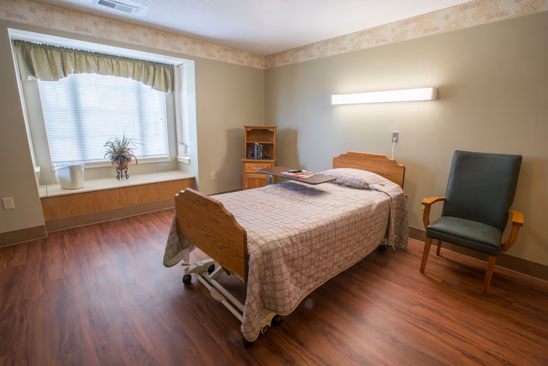 A simple, clean bedroom in a senior living facility with a single bed covered in a checkered bedspread, a green cushioned chair with wooden armrests, a small wooden cabinet, and a large window with blinds and a green valance. The room has wood flooring and a wall-mounted light above the bed.