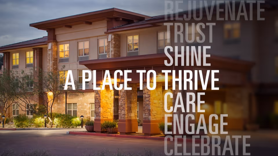 Exterior view of a senior living facility building at dusk with warm lights glowing from the windows and entrance. The building has stone and beige walls with a covered entrance supported by stone columns. Trees and shrubs are visible in front of the building.