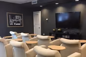Seating area with upholstered armchairs and small wooden side tables facing a wall-mounted flat-screen TV in a dark gray common room.