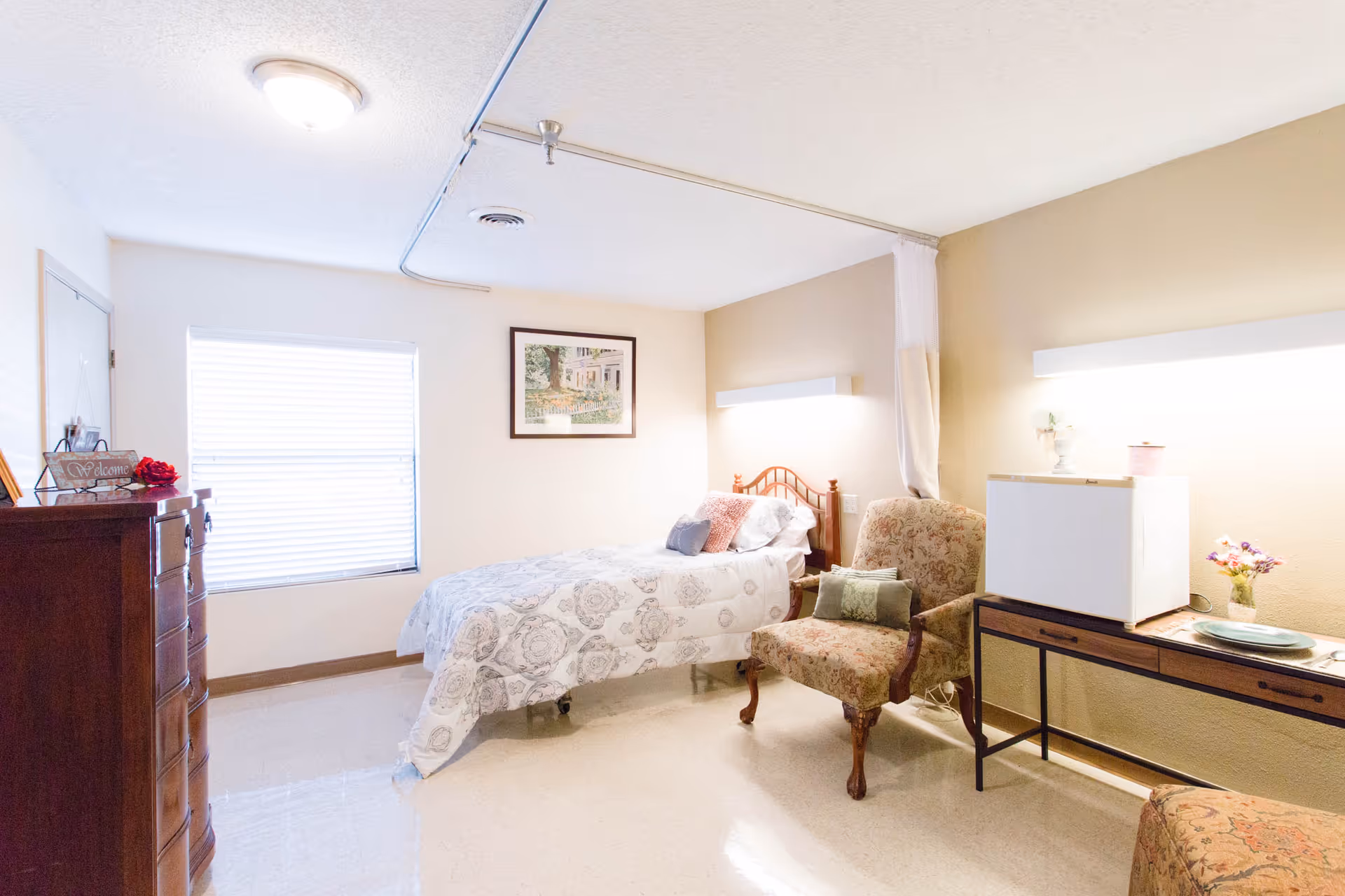A bright and clean bedroom with a single bed covered in a patterned white and gray bedspread. Next to the bed is a floral upholstered armchair with a green pillow. A wooden dresser with a welcome sign and decorative items is on the left side near a window with closed blinds. On the right side, there is a small table holding a mini fridge and a plate, with a small vase of flowers. A framed picture hangs on the wall above the bed, and a ceiling light illuminates the room.