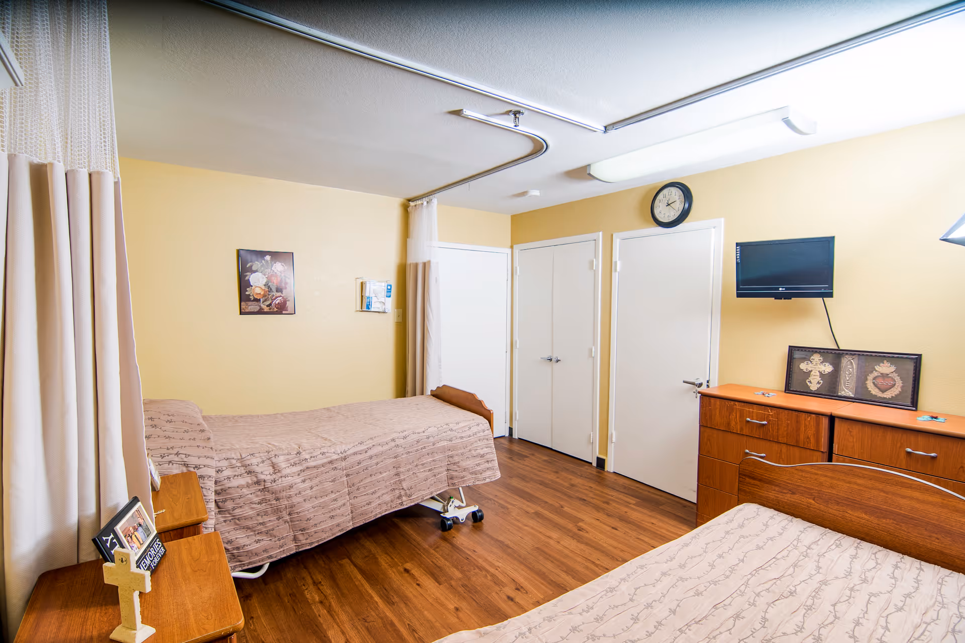 A senior living facility bedroom with two single beds covered in beige patterned bedspreads. The room has wooden flooring, light yellow walls, a wooden dresser with religious artwork and a small wall-mounted TV above it. There is a clock on the wall above two closed white doors. A privacy curtain is partially drawn around one of the beds.