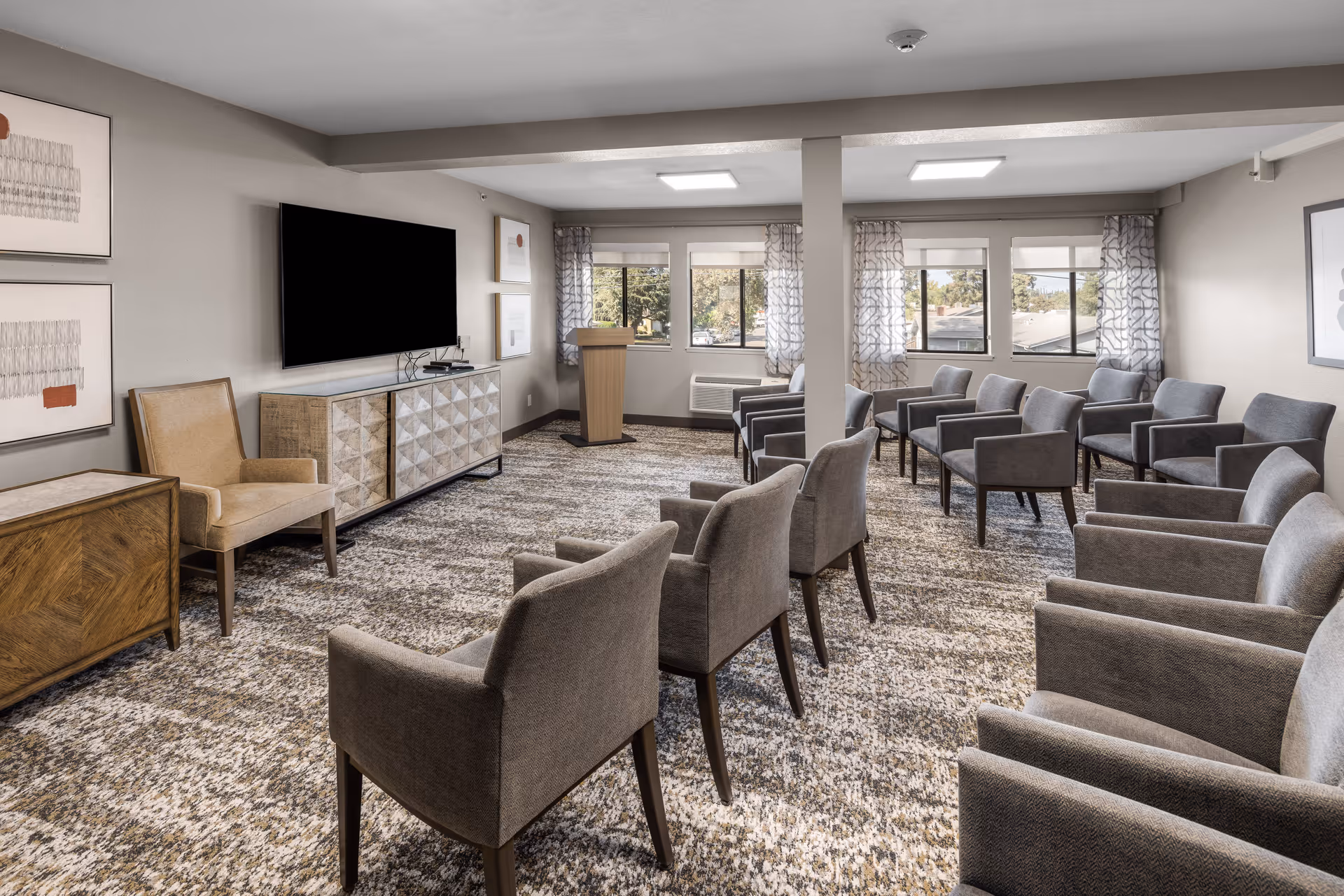 A well-lit meeting or presentation room with multiple gray upholstered chairs arranged in rows facing a large flat-screen TV mounted on the wall. The room has patterned carpet, a wooden podium near the windows with sheer curtains, and framed artwork on the walls.