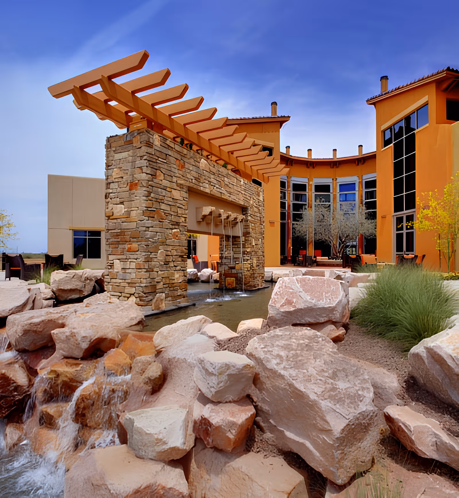 Courtyard with a stone pergola, cascading water feature, and rocky landscaping in front of a curved orange building.